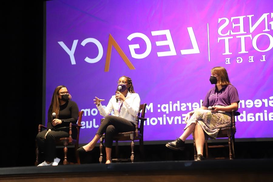 three presenters sit on a stage in front of a screen